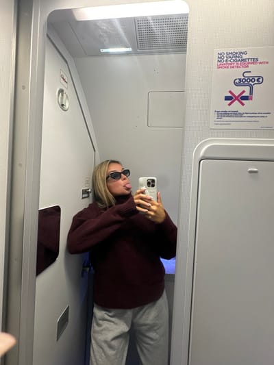 A young woman, wearing a burgundy sweater, grey sweatpants, and black sunglasses, is standing in the bathroom of an airplane, taking a selfie with her smartphone. The bathroom is small and cramped, with white walls and a white door. The woman is sticking her tongue out in the photo. A sign above the toilet is visible, which reads "NO SMOKING NO VAPING NO E-CIGARETTES LAVATORY IS EQUIPPED WITH SMOKE DETECTOR 3000€". The coordinates (latitude: 31.925, longitude: 34.964) place the location in Modi'in Makabim-Re'ut, Israel. The image is taken from the perspective of someone standing outside the bathroom, looking in.