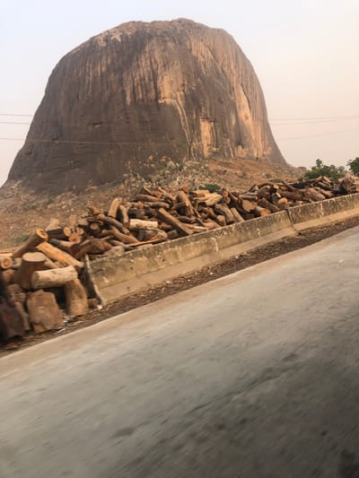 A gray road surface fills the right of the frame. A concrete curb separates the road from a collection of logs piled along the curb. To the left of the wood pile is a large, rocky landform, possibly a mountain or large hill. Some greenery is visible near the top and base of the landform. Thin lines, likely power lines, run from the top of the landform toward the sky. The sky appears hazy or overcast.