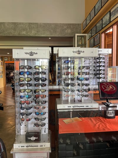 This image depicts the interior of a retail establishment located in the city of Ban Yid, Thailand, showcasing sunglasses. The scene is dominated by two prominent, white and clear acrylic display cases featuring "Ugly Fish" branded eyewear.

The display cases are tall, rectangular structures. Each unit has a white top panel with the "Ugly Fish" logo (a stylized fish head with a helmet-like design) and "Ugly Fish" text in black. The main body of the cases is clear acrylic, allowing full visibility of the merchandise. Numerous pairs of sunglasses with various frame styles and lens colors (including mirrored blue, yellow, red, dark, and light pink) are neatly arranged on internal pegs. Small tags, likely indicating prices or product information, are attached to some sunglasses. The base of the left display case prominently displays the website "www.uglyfisheyewear.com". The right display case shows visible price tags of "1,399.-" and "3,999.-", presumably in Thai Baht.

To the right of the display cases, a counter is present. Its surface is partially covered by a draped orange fabric, upon which a small, gold-colored, folded item (possibly a card or brochure) rests. Below the counter, a neat stack of black boxes, each bearing the "Ugly Fish" logo, is visible. A black monitor on the counter displays the "Ugly Fish" logo in white against a circular red background.

The environment features a light-colored ceiling with a prominent, textured concrete beam running horizontally across the upper portion of the frame. The walls exhibit varying finishes: a section of light-colored wall (possibly off-white or cream) is seen on the left, while a warm yellow or orange wall is visible on the right. This yellow wall features a long, horizontal shelf adorned with multiple framed black-and-white photographs, likely depicting motorcycles or related imagery. Wooden window frames with horizontal slats are partially visible on the far right, suggesting a source of natural light. In the background on the left, a portion of a dark-colored motorcycle, possibly a cruiser, is discernible, along with wooden flooring. A white cabinet containing a gold-colored trophy or decorative item is situated behind the left display case. No individuals are present in the image.
