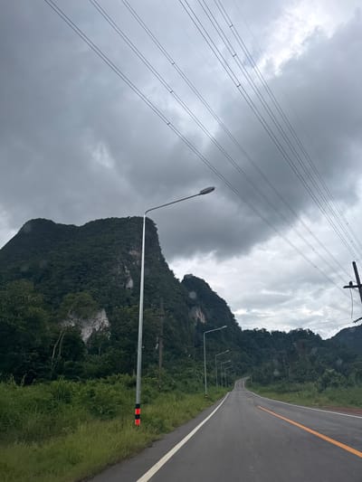 The image depicts a winding asphalt road through a mountainous, forested landscape in Ban Pak Ra, Thailand. The road is marked with a yellow line on the right and a white line on the left, indicating traffic lanes. Along the side of the road, a series of lampposts with arched tops stand in a row, spaced at intervals. One lamppost in the foreground has a red and black striped reflective marker near its base. Overhead, a dense network of power lines crisscrosses the sky, suspended from utility poles. The sky is overcast with heavy, dark clouds, suggesting a day with poor weather conditions, possibly rain. The surrounding mountains are covered in lush, green vegetation, with some rocky outcrops visible on their slopes. The overall scene conveys a sense of isolation and natural beauty, tinged with the somber atmosphere of the cloudy weather. There is no visible text in the image.