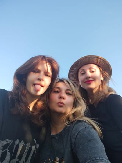 Three young women are taking a selfie outdoors on a clear day. The sky is a bright, solid blue, indicating it is daytime. The woman on the left has long, reddish-brown hair and is sticking her tongue out, her expression playful. She is wearing a dark t-shirt with a graphic design. The woman in the middle has blonde hair and is puckering her lips, also with a playful expression. She is wearing a dark grey t-shirt. The woman on the right is wearing a wide-brimmed straw hat and bright pink lipstick. She has blonde hair and is smiling. She is wearing a black shirt and is also visible from the shoulders up. The lighting suggests it might be late afternoon, as there are warm highlights on the women's faces and hair. The scene appears casual and friendly, with the women posing together for a picture, likely during a social gathering or outing. There is no visible text in the image.