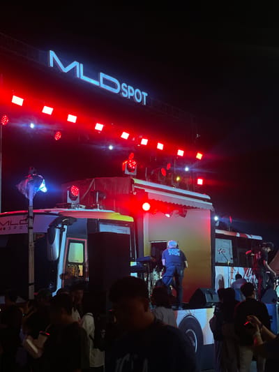 The image captures a live performance at night, likely an outdoor concert or event, in Salembaran Jati, Indonesia. The stage is adorned with the illuminated "MLD SPOT" signage and lined with red stage lights. On stage, a performer in a dark t-shirt and cap can be seen in front of a turntable. The area in front of the stage is packed with attendees. A vehicle with a modified stage on its roof is present, presumably being used for the performance. There are other personnel and equipment situated atop the vehicle. The surrounding environment is dark, highlighting the stage lighting and the performers.