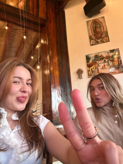 Two women pose for a selfie. One woman is in the foreground, sticking out her tongue. The other woman is in the background, making a kissy face and holding up two fingers in a peace sign. The woman's fingers are adorned with rings. Behind them is a wooden ceiling with string lights and a wall decorated with tiles depicting religious figures and scenes. A black speaker is also visible on the wall. The image is rotated counterclockwise.