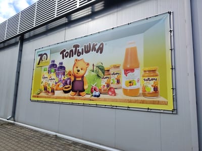 A large, horizontally oriented billboard is mounted on the exterior wall of a light gray, corrugated metal building, likely a commercial structure or warehouse, located in the city of Malaryta, Belarus. The billboard displays a bright, colorful advertisement for "Toptyzhka" brand baby food and juices. The central focus of the advertisement is a cartoon bear character, along with various fruits such as apples, pears, kiwi, raspberries, and strawberries, and several products including jars of puree and bottles of juice. The "Toptyzhka" logo, featuring stylized brown lettering and a small bear cub, is prominently displayed. On the left side of the billboard, a yellow and black graphic marks "70 years" with "1955-2025" and the text "С Днём Рождения!" which translates to "Happy Birthday!". The background of the billboard transitions from a pale blue at the top to a vibrant yellow at the bottom, creating a cheerful and inviting atmosphere. The building itself has a large, modern-looking ventilation or cladding system along the top, with horizontal silver slats. The billboard is attached to the wall with black ties and metal grommets. In the foreground, a paved area with a brick-like pattern is visible, suggesting a pedestrian walkway or parking area. The sky above is partly cloudy.