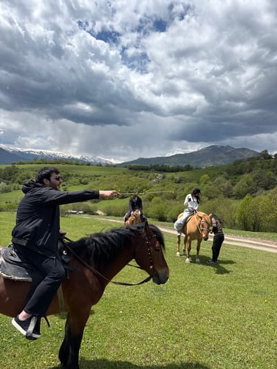 In an outdoor, grassy area of Haghartsin, Armenia, three individuals and two horses are visible. The foreground is dominated by a man on horseback, holding a twig or small plant stem up in the air. He wears a black jacket, pants, sunglasses, and dark shoes. The horse is brown with a black mane. In the background, another horse is ridden by a person in white attire, while another person in darker clothing stands near the horse's head. Further in the distance, mountains with snow-capped peaks and forested hills are visible under a cloudy sky. A dirt path is also present near the second horse.