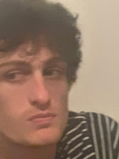 The image presents a close-up, head-and-shoulders view of a single male individual. The location is specified as Roanne, France, though no specific geographical markers within the city are visually present in the image. The scene is indoors.

**Subject:** A young adult male with fair skin and dark, curly hair. His eyes are dark, with the left eye (viewer's left) directed slightly to the side and the right eye (viewer's right) looking forward or slightly right. His nose is prominent. His mouth is closed, exhibiting a neutral to slightly pensive expression. He is wearing a dark garment featuring multiple light-colored diagonal stripes across the chest and shoulder area.

**Environment:** The background consists of a plain, light-toned wall, possibly off-white or beige, providing no further contextual details. The lighting suggests an interior setting with no visible external light sources. No other individuals or objects are present within the frame.

**Actions/Interactions:** The individual is static, posing for the image. No active actions or interactions with other subjects or objects are observable.