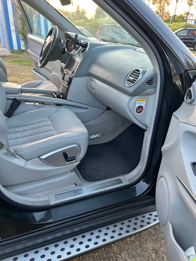 The image shows the interior of a Mercedes-Benz SUV, viewed from the exterior passenger side with the door open. The driver's seat, made of light grey leather and fabric, is prominently featured. The dashboard and steering wheel are visible, as is the center console with various controls and a media display. A phone mount is attached to the steering column. A warning sticker with an airbag symbol is affixed to the lower dashboard. A black rubber floor mat is on the floor, and a mesh storage net is visible behind the driver's seat. The Mercedes-Benz logo is visible on the door sill. Outside the vehicle, dry grass and a blurred background suggest an outdoor setting, possibly a parking area. The lighting indicates it is daytime, with some sunlight filtering through. The scene is devoid of any people or activity. The image appears to be taken in Grao, Spain, based on external information not visible in the image itself.