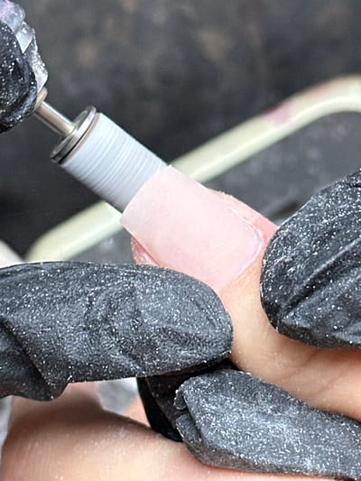 The scene, located in Kafr Kasim, Israel, depicts a close-up perspective of a nail care procedure. A gloved hand, adorned with a dark, textured glove coated in fine, light-colored dust particles, is operating an electric nail file. The file features a silver metallic shaft holding a gray, cylindrical abrasive sanding band, which is actively engaged in shaping the surface of an artificial nail. This nail, appearing light pink and opaque, is applied to a human finger, which is partially visible. Another hand, also wearing a dark, textured glove and similarly coated in dust, is partially visible supporting or stabilizing the finger and nail being worked on. The immediate background is dark and indistinct, consistent with an indoor setting.
