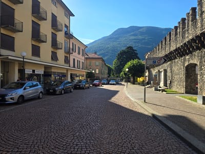 The image depicts a sun-drenched street in Bellinzona, Switzerland, featuring a view towards the Castel Grande. On the left, a pale yellow building with balconies and shops lines the street, displaying signs such as "HOOKAH" and "VAPE SHOP." Several cars are parked along the cobblestone street. A silver hatchback is parked closest to the foreground, followed by a dark gray sedan and a red sedan. Further down the street, more cars are visible, some turning a corner.
The right side of the street is dominated by a historic stone wall, characteristic of Bellinzona's castles, topped with battlements. A street sign indicating "Polizia - Municipio" (Police - Municipality) with a parking symbol and a speed limit of 2.5 km/h is attached to this wall. Beyond the wall, trees and the slopes of a forested mountain rise under a clear blue sky. The street itself is paved with patterned cobblestones, with a sidewalk running alongside the stone wall. Sunlight casts strong shadows across the road, indicating it is likely late morning or early afternoon. A single lamppost stands near the shops. A few individuals are visible, one near the shops and another walking further down the street. The overall impression is a picturesque European town scene with a blend of historical architecture and modern life.