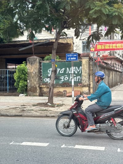 A man wearing a blue shirt and grey pants rides a black and red motorcycle in Nam Đông Hà Ward, Vietnam. He wears a blue helmet and light blue flip-flops. The motorcycle has "YAMAHA" written on the side. In the background, there is a weathered green wall with white graffiti. A blue street sign that reads "KIỆT 290 LÊ DUẨN" is visible. A red banner with Vietnamese text hangs from a building. Trees with dense foliage overhang the street. The road surface is asphalt with white painted markings.