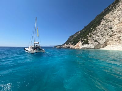 The image shows a bright, sunny day with a clear blue sky over a calm, turquoise sea. On the left side of the frame, a white catamaran sailboat with a mast and yellow flag is anchored. Several people are visible on the deck of the boat. The water around the boat is crystal clear, revealing the sandy bottom in some areas.

To the right of the frame, a steep, rocky cliff face rises from the sea, covered in patches of green vegetation. A small white sandy beach is visible at the base of the cliff. In the distance, other small boats can be seen on the horizon, and a peninsula with more cliffs and vegetation extends into the sea. The overall impression is one of a peaceful, idyllic coastal landscape, likely a popular spot for sailing and swimming. The location is near Platrithias, Greece, suggesting a Mediterranean setting.