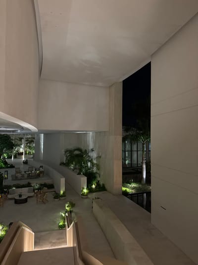 A contemporary courtyard is captured from an elevated perspective at night. Clean white walls frame the scene. A glimpse of a dark, open-air space with trees can be seen above. Below, a curated garden with lush greenery and strategically placed lighting is visible. A seating area with light-colored furniture sits to the bottom of the image. The overall aesthetic is modern and minimalist.