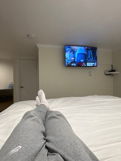 A person wearing grey sweatpants and white socks lies on a white bed in a room with a television mounted on the wall. The television is playing content in Bulgarian. The room appears to be a hotel room. The location is in Hampton, United States of America.