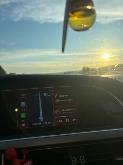 The image captures a dashboard view from inside a car, with the sun setting outside. A yellow, translucent object, possibly a charm or air freshener, hangs from the windshield, reflecting the sunlight. The car's infotainment screen displays navigation and music information. The navigation shows a route from "Egoot" to Sofia, with an estimated time of arrival of 21:57. The current speed is indicated as 64 km/h. The music app is showing "Remember t..." by Michael Jackson. Below the navigation, the time is displayed as 19:59, with a 5G signal. The weather appears clear with a bright, hazy sky due to the setting sun. The scene suggests a journey taking place in the late afternoon or early evening. The location is identified as Ignatievo, Bulgaria.