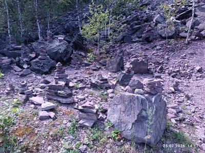 The image depicts an outdoor scene in a rocky, wooded area, possibly a quarry or a natural rock formation. There are numerous cairns, which are stacks of stones, of various sizes and heights scattered throughout the foreground and midground. The stones are predominantly gray and reddish-brown. In the background, a dense growth of trees with slender white trunks and green leaves lines the upper portion of the image, creating a backdrop of foliage and darker rocks. The ground is covered in loose stones and some sparse vegetation, including green plants and moss. The lighting suggests it is daytime, with a slightly overcast or subdued light. A date stamp "25.07.2026 14:17" is visible in the lower right corner. There are no people or animals visible in the image. The environment appears rugged and natural, with the cairns suggesting human presence and activity, possibly for marking trails or as a form of art. The location is identified as Assy, Russia.