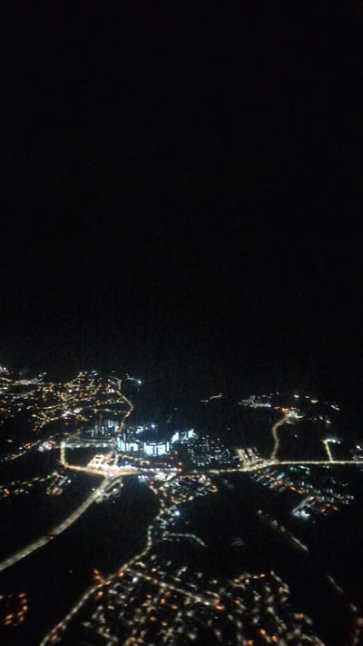 This is an aerial photograph taken at night, depicting a coastal urban landscape, likely Port Dickson, Malaysia. The image is dominated by the stark contrast between the dark expanse of the sea on the left and the illuminated landmass on the right.
**Subjects & Scene:** The main subjects are the artificial lights of a city, viewed from above. These lights outline various urban features such as roads, buildings, and possibly industrial areas or port facilities. The scene is a vibrant nighttime city, with a clear boundary marking the coastline.
**Activity or Event:** The image captures the static appearance of a city illuminated after dark. There is no discernible specific activity or event taking place, but rather the general state of an active urban environment at night.
**Notable Details:**
* **Time of day:** Clearly night, indicated by the black sky and the sole reliance on artificial lights for visibility.
* **Weather:** The night appears clear, with no visible clouds or atmospheric interference.
* **Location cues:** The distinct line of lights along the edge suggests a coastline. The patterns of lights include bright clusters indicating dense development, linear streaks representing roads, and some dispersed lights in less populated areas. Some areas show a grid-like pattern of streets, while others appear more organically developed along the coast. The overall appearance is consistent with an illuminated coastal city.
* **Emotions:** Not applicable.
* **Visible text:** There is no visible text in the image.
The illumination ranges from bright white and yellow lights to more diffused glows, showcasing the varied light sources within the city. The dark area to the left is vast and featureless, strongly suggesting open water.