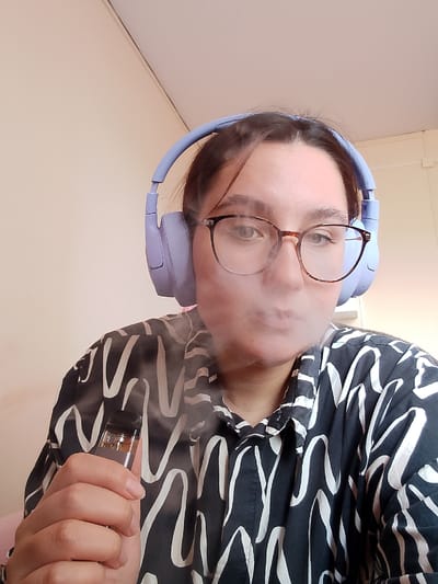 A medium shot shows a young woman with brown hair, wearing light purple headphones and glasses with brown rims. She is exhaling a cloud of vapor, holding a vape pen in her right hand. She is wearing a black shirt with a white wavy pattern. The background is a plain, light-colored wall. There is no indication of the city of Birsk, Russia.