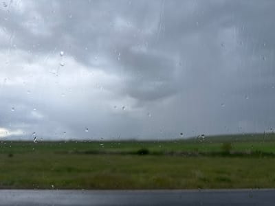 The image shows a rainy view through a window. The sky is overcast with gray clouds. In the background, there is a green field. A dark gray or black surface, likely a road, is visible at the bottom of the frame. Raindrops are present on the window. The location is unknown.