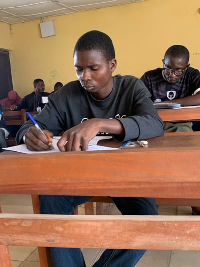 The image depicts a classroom scene in Abobo, Nigeria, focusing on a young adult male seated at a light brown wooden desk in the foreground. This individual, with a dark complexion and short dark hair, wears a dark grey long-sleeved sweatshirt and blue jeans. He is actively writing on a white sheet of paper with a blue ballpoint pen held in his right hand, while his left hand rests on the paper. His gaze is directed downwards at the task, indicating concentration. An eraser and a clear plastic ruler are also visible on his desk surface.
In the middle ground to the right, another young adult male with glasses is seated at a similar wooden desk, looking down at papers. He wears a dark t-shirt with a white graphic. Additional individuals are partially visible in the background, including one male in a dark jacket with a lanyard and another in a red hooded garment, all appearing to be seated and focused on tasks.
The classroom environment features pale yellow walls, some displaying faint markings and what appears to be mathematical notation. A white tiled ceiling and a white rectangular fixture are visible on the upper left wall. The floor is light-colored. The scene suggests a formal academic setting, such as an examination or study session.