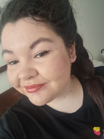 A medium shot shows a young woman with dark hair and brown eyes, wearing black clothing. She has makeup on, including eyeliner and lipstick. The background is blurry and appears to be indoors, with a hint of furniture and a wall. The image is taken from a slight angle, looking up at the woman. No specific location within Carlisle, United Kingdom, can be identified from the image.