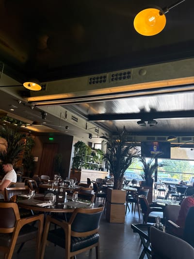 This image captures the interior of a modern restaurant or cafe identified as "CONCEPT," located in Strumica, North Macedonia. The perspective is from a seated position, looking across the dining area towards large windows that offer an outdoor view.
The establishment features a dark ceiling, likely black or dark grey, from which industrial-style pendant lamps with yellow/orange shades cast warm light. Sections of the ceiling incorporate olive-green elements with visible ventilation grilles and circular fixtures, alongside multiple small, white dome-shaped surveillance cameras. An illuminated green "EXIT" sign is positioned above a wooden door in the mid-left background.
The dining area is furnished with numerous tables, primarily circular, featuring dark, reflective surfaces (likely glass) and light-toned wooden bases. These are paired with chairs that have light wooden frames and dark, possibly black or dark brown, upholstered seats and backs. Several tables are set with clear, stemless drinking glasses and dark placemats. One table in the central foreground displays three glasses and two small black menu holders.
Prominent tropical plants, including large potted palm trees, are strategically placed throughout the interior, providing natural divisions and decor. A wall on the right side of the frame features shelves holding multiple smaller potted green plants.
Two individuals are clearly visible within the establishment. In the mid-left foreground, a male with short, dark hair is seated at a table, facing right, wearing a white short-sleeved top. His left arm rests on the table. In the mid-right, another person is partially visible from behind, wearing a red long-sleeved top, seated at a table and facing left.
Large windows along the far wall reveal an outdoor scene. This view includes several dark-colored vehicles, indicative of a street or parking area, and distant green foliage or hills under a bright sky. Centrally positioned above the windows is a large flat-screen display showing a glowing logo and the word "CONCEPT" in white text. Black speaker units are mounted on the ceiling structure near this display. The overall ambiance is characterized by a blend of artificial warm lighting and natural light from outside, creating a functional and contemporary dining environment.
