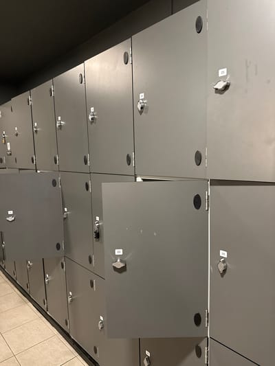 The image displays a section of grey metal lockers arranged in a grid. The lockers have round, black knobs or push-button locks. Some locker doors are ajar, revealing the interior. Several lockers are labeled with white rectangular stickers displaying numbers, such as 14, 22, 23, 25, and 26. The lockers are positioned in a room with a dark ceiling and tiled flooring. The perspective suggests an indoor environment, likely a public facility such as a gym or changing room. There are no people visible in the image. The overall scene is orderly and functional. This image does not provide any information about its location in Carlow, Ireland.