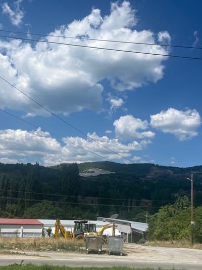 A wide shot captures a sunny day with fluffy white clouds against a bright blue sky. In the foreground, two yellow backhoe loaders are parked next to a large, metallic dumpster. Behind them, a building with a red roof and white walls is visible, with text that reads "BELLMONDO The Shoe House." A gentle slope rises in the background, covered in dense, dark green trees. Power lines crisscross the upper portion of the image. The overall scene appears to be an outdoor industrial or construction area.