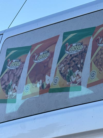 The image shows a close-up of a white van's window in Shumen, Bulgaria, covered with a perforated vinyl sticker displaying advertisements for nuts. The sticker features multiple repeating panels, each showcasing a product image of nuts, likely almonds, walnuts, and pistachios, arranged in a visually appealing manner. A stylized squirrel logo and the brand name "Unezy" are prominently displayed in the center of each panel, accompanied by smaller text in what appears to be Cyrillic script, possibly indicating the type of nuts or a slogan. The sticker is adhered to the exterior of the van's window, and the texture of the vinyl is visible as a fine grid pattern. The background outside the window is a clear blue sky, with a thin green wire, possibly an antenna or power line, running diagonally across the upper left portion of the image. The van's white paint is visible along the bottom edge of the frame, showing some minor scuffs. The overall scene suggests a commercial vehicle used for advertising or delivery services in the city.