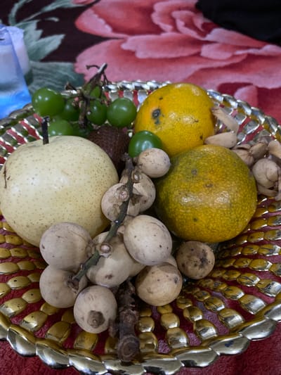 In the city of Seuneubok, Indonesia, a decorative gold-colored platter holds a variety of fruits. The platter is positioned on a red, floral-patterned fabric. The fruits include a large, pale yellow pear with a stem, a cluster of small green grapes, two oranges, a dark, spiky fruit that appears to be a salak, and a bunch of small, pale, round fruits on stems, possibly longkong. Some dried, light-brown seed pods are also present in the platter. The lighting suggests an indoor setting.