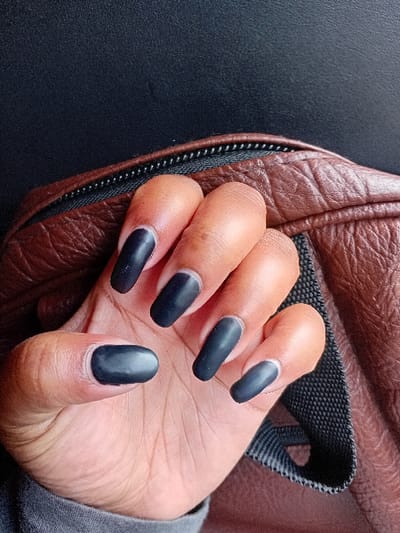 The scene depicts a close-up of a human left hand, exhibiting a brown skin tone, located in Mururi, Kenya. The hand features five fingernails (thumb, index, middle, ring, pinky) which are long, oval-shaped, and coated with a matte black or very dark grey polish. A subtle light outline is present at the base of each nail, just above the cuticle. The hand is positioned resting on a textured brown material, consistent with leather or synthetic leather, which includes a black zipper with white teeth. A black woven textile strap is visible underneath the fingers. The lower part of the hand is covered by a grey fabric, consistent with a sleeve. The immediate background is an indistinct dark surface.