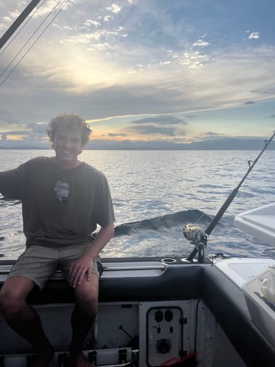The image depicts a single male subject seated on the stern of a recreational fishing boat on open water off the coast of Port Kembla, Australia. The scene is illuminated by the low angle of the sun, suggesting either sunrise or sunset, with light reflecting off the water and scattered clouds in the sky.

The male, appearing to be a young adult, has light-colored curly hair, is barefoot, and wears a dark olive-green short-sleeved t-shirt with a light-colored graphic and light-tan shorts. He is positioned facing forward and slightly to the left, looking directly at the camera with a smile. His left arm is partially raised, while his right hand rests on his right thigh.

The boat features a dark-colored padded coaming along the stern, with two integrated black cup holders. A light-colored panel containing multiple switches and a circular dial is visible below the seating area. On the starboard side of the stern, a heavy-duty fishing rod equipped with a large gold-colored conventional reel is secured in a rod holder, angled outward, with a fishing line extending into the water. The boat is actively moving, indicated by the wake visible on the water surface directly behind the vessel. A white cooler or bait tank is partially visible on the far right.

In the background, the distant coastline of Port Kembla is discernible, featuring industrial structures including chimneys or stacks, consistent with the Port Kembla steelworks. Beyond these structures, a range of mountains or hills is present, silhouetted against the horizon under a partially cloudy sky.