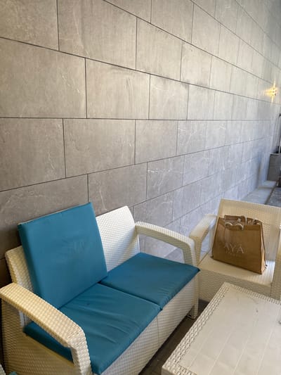 The image depicts an outdoor seating area with white wicker furniture against a grey tiled wall. The furniture includes a sofa with bright blue cushions and a separate armchair. A brown shopping bag with the "AYA RESORT" logo is placed on the armchair. A white coffee table is situated in front of the sofa and armchair. The wall behind the furniture is constructed of large, rectangular grey tiles with subtle marbling. The overall scene suggests a relaxed outdoor setting, possibly at a resort or a similar establishment. There are no people visible in the image. The lighting suggests it is daytime.