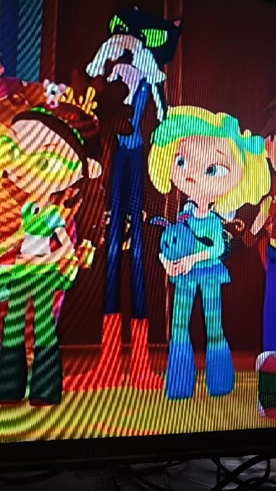The image displays a photograph of a television or monitor screen, exhibiting prominent diagonal scan lines, located in the city of Kasansay, Uzbekistan. The screen shows an animated scene featuring four cartoon characters within an indoor environment characterized by dark red walls and a horizontally striped floor with yellow and orange tones.
From left to right:
1. A bipedal character, predominantly green with white and red stripes, wearing a multi-colored hat adorned with brown antler-like structures. Its left arm is slightly raised.
2. A tall, slender character in dark blue clothing and red boots. A small white creature, resembling a mouse or ferret, is positioned at its chest. Above this character's head, a black cat-like silhouette with glowing green eyes is visible.
3. A blonde-haired girl, dressed in a light blue-green striped jumpsuit, holds a small blue animal, possibly a rabbit or plush toy, close to her chest. Her facial expression conveys concern or surprise, with wide eyes and a slightly open mouth.
4. A partially visible character on the far right, showing a segment of red and green attire.
The characters are standing, appearing to observe or react to an unseen event within the animated narrative.