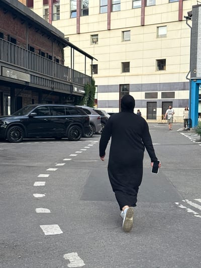 A person wearing a black abaya and white sneakers walks away from the camera on an asphalt road. They are holding a white smartphone in their right hand. Dashed white lines are painted on the road, marking a lane or pathway. In the background, two black SUVs are parked, and behind them are buildings. The building on the left has a brick facade and a dark railing on an upper level. The building on the right is a multi-story structure with a cream-colored facade and square windows. Further in the distance, other people are visible walking. The scene appears to be in an urban environment, possibly a street or courtyard in Zhabkino, Russia.