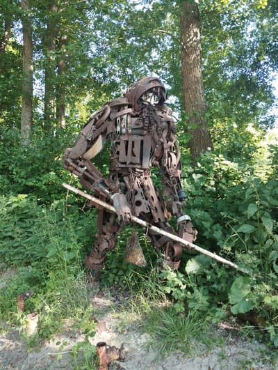 The image shows a full-body, life-sized sculpture of a humanoid figure made from scrap metal. The figure is standing in a lush, green outdoor setting, surrounded by dense foliage and trees. The sculpture appears to be rusted, giving it a weathered and aged look. It is posed as if holding a long, thin object, possibly a spear or staff, with its right arm. The figure's head is covered by a helmet-like structure, and parts of its body are constructed from various metal components such as gears, chains, and plates.

The scene is set during the daytime, with sunlight filtering through the trees, casting dappled light on the sculpture and the surrounding vegetation. The overall impression is one of a somewhat imposing, perhaps even ancient, warrior or sentinel guarding the natural environment. The ground beneath the sculpture is a mix of dirt and gravel, with some smaller pieces of rusted metal visible. The thick green leaves and tall grass suggest a temperate climate and a possibly rural or park-like location. There is no visible text in the image.