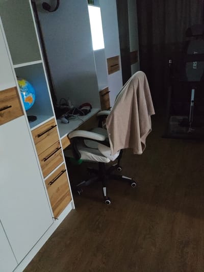 The image depicts an indoor scene, likely a room in a home or office. In the foreground, a white and grey gaming chair with a light brown blanket draped over its headrest is positioned on a dark brown carpeted floor. The chair has black casters and armrests. To the left of the chair is a white and wood-grain cabinet with several drawers. On the top shelf of the cabinet, a small globe with blue and orange continents is visible. Behind the chair, a desk is partially visible, with some electronic devices and headphones on its surface. The background is dimly lit, with a large, bright rectangular light source on the wall. To the right of the chair, a dark exercise machine with the word "ERS T" visible on it is present. There are no people visible in the image. The scene appears to be set in Arshaly, Kazakhstan, based on the provided location information, though no specific visual cues in the image confirm this.