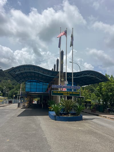 This media file captures a view of the Wang Kelian Immigration, Customs, Quarantine, and Security (ICQS) complex in Perlis, Malaysia. The scene is dominated by the architectural structure of the border crossing, featuring a large, curved metal roof supported by a steel framework, with a blue-tinted glass facade. The area appears to be a road leading into the complex, with an asphalt surface and a raised blue planter with lush green plants in the foreground.
Several flags are visible, including the Malaysian flag, which is striped red and white with a blue canton containing a yellow crescent moon and star. Other flags, appearing purple and green, are also flying.
A prominent sign displays text in both Malay and English. The Malay text reads "JABATAN IMIIGRESEN MALAYSIA" and "POS IMIIGRESEN WANG KELIAN PERLIS". The English translation is likely "Malaysian Immigration Department" and "Wang Kelian Immigration Post, Perlis".
The sky is partly cloudy with dramatic white clouds against a pale blue background, suggesting it is daytime. Lush green trees and foliage are visible behind the complex, indicating a natural, possibly hilly or mountainous, environment. The overall impression is a functional border crossing point under a bright sky, set within a tropical landscape. No specific activity or people are clearly discernible, giving the image a static, observational feel. The image is oriented vertically, with the sky at the top and the road at the bottom.