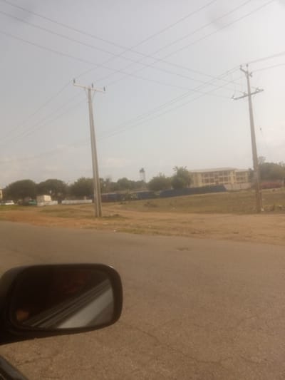 The image depicts a street scene in Kinkinau, Nigeria, under an overcast sky. In the foreground, a car's side mirror is visible, reflecting a portion of the road and possibly the vehicle's interior. The road itself is paved and appears cracked in places. Beyond the road, a grassy area stretches towards a building in the background. This building is a light-colored, multi-story structure with visible windows. Power lines crisscross the sky, supported by utility poles. Several trees are scattered throughout the landscape. A water tower can be seen in the distance on the left side of the image. Dark blue tarps are spread out in the middle ground. The scene suggests an urban or semi-urban environment within Kinkinau.