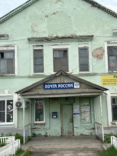 The image shows the Ленино, Russia Post Office, a two-story pale green building with a peaked wooden overhang above the entrance, displaying a sign that reads "ПОЧТА РОССИИ" in Cyrillic. The paint on the building is peeling, and there are signs of weathering and age. The windows on the building are rectangular with dark frames and shutters. There are signs and posters on the building's exterior. The entrance is situated on the ground floor, with a concrete walkway leading to the doorway. In the lower windows, there are decorations related to Victory Day, including Soviet symbols. There are also utility boxes and AC on the lower right.
