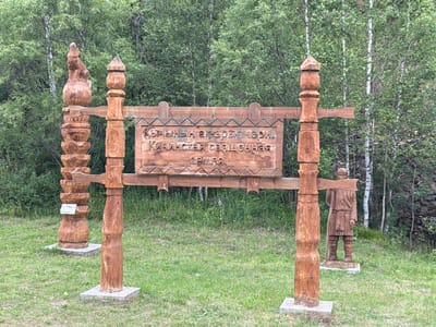 The image shows a wooden monument in a grassy area, likely in Srednie Kichi, Russia. The monument features carved wooden posts supporting a wooden sign. The sign has text carved into it, with one line appearing to be in Cyrillic and another in a different script, possibly Altai. To the left of the sign is a tall, carved wooden post topped with a figure that resembles a wolf howling. To the right of the sign is another wooden post, and further to the right, a carved wooden figure of a person in traditional clothing stands on a small pedestal. The background consists of lush green trees, with some birch trees visible. The overall scene suggests a cultural or historical site within a natural setting.