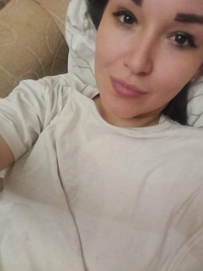 The media file is a close-up, selfie-style photograph of a woman lying down. She is wearing a light-colored long-sleeved shirt. Her face is partially visible, with her eyes and mouth in focus. She has dark hair and appears to be wearing makeup. The background consists of a light-colored textured surface, possibly a sofa or bed, and some crumpled bedding with a subtle pattern. The lighting suggests it could be indoors, potentially during the day or evening. There is no visible text in the image. The overall impression is a casual, relaxed moment.