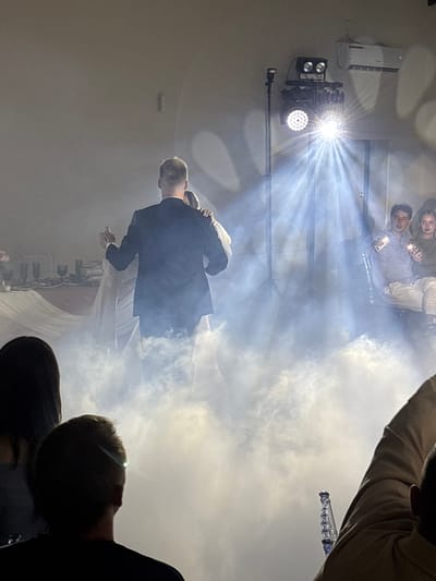 In Нижние Муллы, Russia, a wedding reception is underway. The image captures a newlywed couple in the foreground, engaged in their first dance. They are enveloped in a cloud of fog, illuminated by dramatic spotlights. To the right, a group of guests is seated, observing the dance. The background features a table laden with dishes, suggesting a formal dining setup. The overall ambiance is festive and romantic, enhanced by the strategic use of lighting and fog effects to create a magical atmosphere.