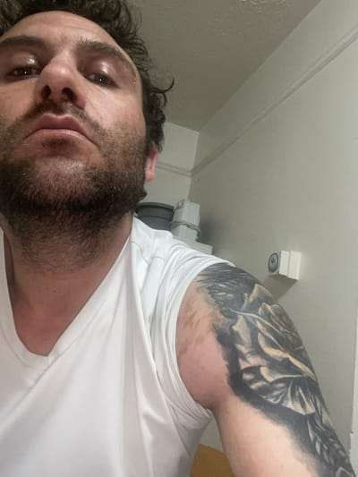The image shows a close-up, low-angle shot of a man's face and upper body, taken indoors. The man has curly dark hair, a beard, and appears to be wearing a white t-shirt. He has a tattoo on his right arm, which appears to be a large floral design. The background consists of a neutral-colored wall with some shelving and objects visible, suggesting a domestic or utility space. There is no indication of the city of Armagh, United Kingdom, or any other specific location.