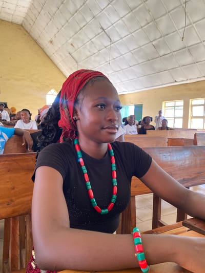 The media file depicts a gathering of people in what appears to be a community hall or church in Dutsen Kato, Nigeria. The image is oriented vertically, with the primary subject, a young woman, prominently featured in the foreground. She is wearing a black top adorned with subtle sparkle details, a red headscarf, and a beaded necklace and bracelet in alternating red and green colors. Her gaze is directed slightly upwards and to the left, with a neutral expression.
Behind her, a row of other individuals are seated at wooden benches. Some are looking towards the camera, while others appear to be engaged in conversation or observing something. The setting suggests an indoor event, possibly a service or a community meeting, given the presence of multiple people seated in rows. The room has a high, white, textured ceiling and yellow walls. Windows with visible panes are present, suggesting natural light is entering the space, though the time of day is not explicitly discernible.
One individual in the background, wearing a white t-shirt, has text on it, but it is partially obscured and unreadable. Another person in a white garment is visible further back. The overall atmosphere seems calm and communal. The wooden furniture and ceiling design are consistent with local architecture or community building styles.