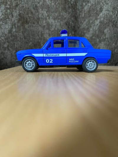 A blue toy police car with white racing stripes and the word "ПОЛИЦИЯ" (POLITSIYA) in white lettering is the subject of the image. The car also has the number "02" and "МВД РОССИИ" (MVD ROSSII) in white lettering. It is positioned on a wooden surface, with a textured brown backdrop. The car has a blue siren light on its roof. The scene is indoors. The location of Бескөл ауылдық округі, Kazakhstan is not depicted in this image.
