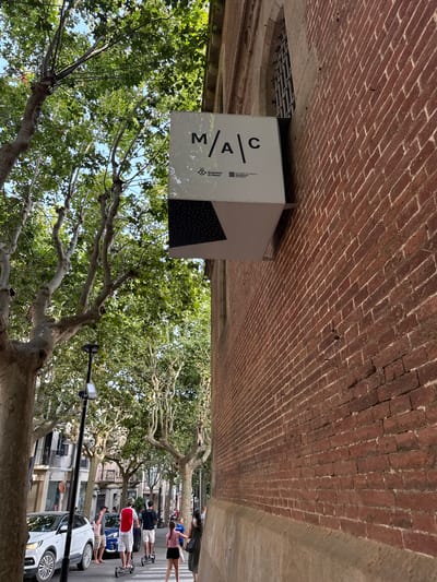 The image shows a sign for "MAC" which stands for "Museu d'Art Contemporani de Barcelona" (Museum of Contemporary Art of Barcelona) on a red brick building in Mataró, Spain. The sign is white with black text and a logo. The building has a arched window with a barred grille. To the left of the building, there are trees with green leaves, a street lamp, a white car, and several people walking and riding electric scooters. The street is lined with trees and buildings. The sky is visible and appears to be blue with some clouds.