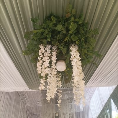 A detailed view of a decorated ceiling in Pasir Muncang, Indonesia, is presented. A large, central fixture is suspended overhead, composed of a dense cluster of artificial green foliage. Cascading from this foliage are multiple long strands of artificial white flowers. A prominent white, spherical light fixture is embedded within the green foliage and white flower arrangement. The ceiling itself is covered with draped and pleated fabric, exhibiting alternating sections of light green and white material. No individuals, actions, or interactions are present in the frame.