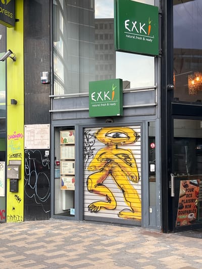 This image depicts the exterior of an EXKi restaurant in Saint-Josse-ten-Noode - Sint-Joost-ten-Node, Belgium. The facade features a bright green EXKi sign with a stylized carrot logo and the tagline "natural, fresh & ready." To the left of the entrance, a security camera is mounted on a lime green pillar. The storefront entrance is partially covered by a roll-up metal gate, which has been adorned with graffiti of a yellow, alien-like figure with a large eye and a stylized body. Inside the visible portion of the storefront, several shelves are stocked with food products. To the right of the entrance, another EXKi sign displays food items with the text "POUR DES PLAFIRS NON COUPABLES." The ground in front of the building is paved with a patterned brick walkway. The overall scene suggests an urban environment with commercial activity.