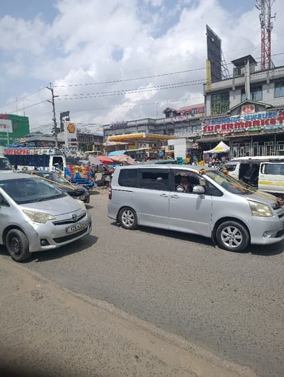 The image displays a busy street scene under an overcast sky in Matungulu west ward, Kenya. In the foreground left, a silver Toyota Ractis with Kenyan license plate KCX 331T is partially visible, featuring a dark-colored steel rim on its front left wheel and minor scuffs on the front bumper. Centrally, a silver Toyota Noah minivan, decorated with white and yellow ribbons/flowers on its hood and rearview mirrors, is in motion, with a driver discernible within. In the background, a Shell gas station with yellow and red branding, including fuel pumps and canopies, is visible. To the right, a multi-story building housing "MUMUNI SUPERMARKET" with a prominent red and blue sign, along with a cellular tower, is present. Market stalls with various canopies, including white and red, are set up between the main road and the buildings. Further into the background, a colorful, decorated truck and other vehicles are partially visible. Utility poles and overhead power lines traverse the upper portion of the frame, above the paved asphalt road surface.