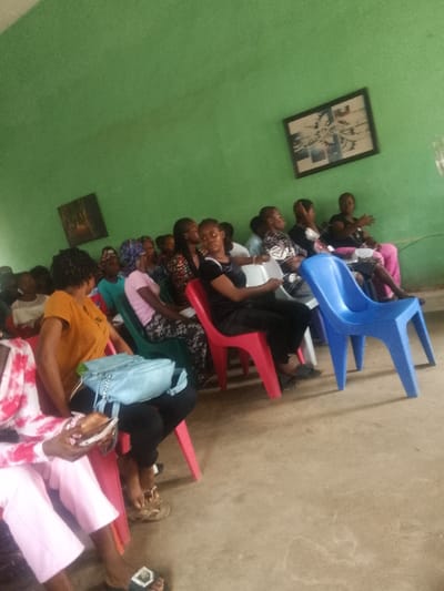 An indoor setting in Umuokpu, Nigeria, depicts approximately 15-20 individuals, primarily women and young women, seated in rows of various colored plastic chairs (red, blue, green, white) on a light-colored concrete floor. The walls are painted a uniform light green. Two framed pictures are mounted on the far wall; a larger, rectangular frame containing an abstract design is located in the upper right, and a smaller, square-ish frame is positioned to its left. Individuals are dressed in casual attire, including t-shirts, patterned blouses, and headscarves. Several attendees hold white papers or booklets. A woman in the midground, wearing a black t-shirt, directly faces the camera. In the foreground, a light blue purse rests on a person's lap, and another individual holds a small, light-colored object. The assembled group suggests an audience attending a meeting or presentation.